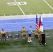 A military ceremony is taking place on the field of an indoor football stadium near the 50-yard line. A uniformed soldier is shaking hands with a civilian at a podium flanked by flags, while several other military personnel in camouflage uniforms are seated in folding chairs applauding. The setting suggests a formal recognition or award ceremony conducted during a sporting event.