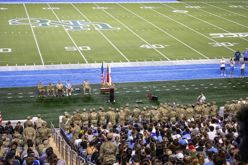 A military ceremony is taking place on the field of an indoor football stadium, with a large formation of soldiers in camouflage uniforms standing at attention near the sideline. A speaker addresses the gathering from a podium flanked by the American flag and what appears to be a state flag, with several seated officials nearby. The stadium seating is filled with a large civilian audience observing the event.
