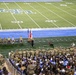 A military ceremony is taking place on the field of an indoor football stadium, with a large formation of soldiers in camouflage uniforms standing at attention near the sideline. A speaker addresses the gathering from a podium flanked by the American flag and what appears to be a state flag, with several seated officials nearby. The stadium seating is filled with a large civilian audience observing the event.