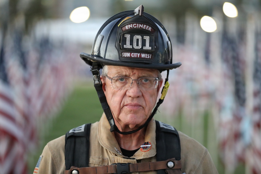 An older male firefighter wearing a black helmet labeled 'Engineer 101 Sun City West' stands in front of a field of small American flags. He is dressed in full firefighting gear including a tan turnout coat and harness straps. His expression is solemn and serious, suggesting a commemorative or memorial context.