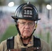 An older male firefighter wearing a black helmet labeled 'Engineer 101 Sun City West' stands in front of a field of small American flags. He is dressed in full firefighting gear including a tan turnout coat and harness straps. His expression is solemn and serious, suggesting a commemorative or memorial context.