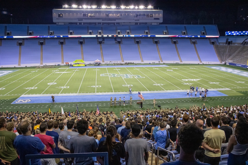 A large crowd has gathered on the field and in the lower stands of a college football stadium at night for what appears to be a military ceremony or patriotic event. Several uniformed military personnel are visible on the field near an American flag, with a podium present suggesting a formal address. The stadium is identified by visible signage as Kenan Stadium, home of the UNC Tar Heels, featuring the characteristic light blue seating.