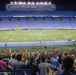 A large crowd has gathered on the field and in the lower stands of a college football stadium at night for what appears to be a military ceremony or patriotic event. Several uniformed military personnel are visible on the field near an American flag, with a podium present suggesting a formal address. The stadium is identified by visible signage as Kenan Stadium, home of the UNC Tar Heels, featuring the characteristic light blue seating.