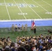 A military ceremony or formal event is taking place on an indoor football field, with a speaker at a podium flanked by the American flag and another flag. A large crowd of attendees, many in military camouflage uniforms, stand facing the field in what appears to be a moment of solemnity or prayer. The football field markings, including the 40 and 50-yard lines, are clearly visible along with a logo on the field.