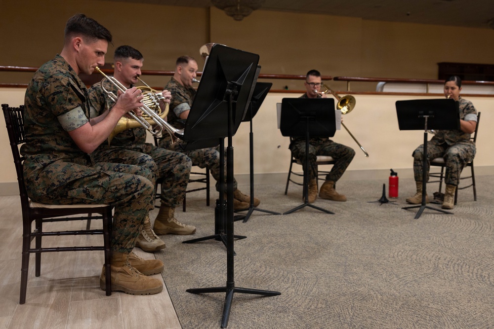 A group of military personnel in Marine Corps digital camouflage uniforms are seated in chairs performing as a brass ensemble. They are playing brass instruments while reading sheet music from black music stands. The setting appears to be an indoor rehearsal or performance space with a carpeted floor and tan walls.