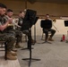 A group of military personnel in Marine Corps digital camouflage uniforms are seated in chairs performing as a brass ensemble. They are playing brass instruments while reading sheet music from black music stands. The setting appears to be an indoor rehearsal or performance space with a carpeted floor and tan walls.