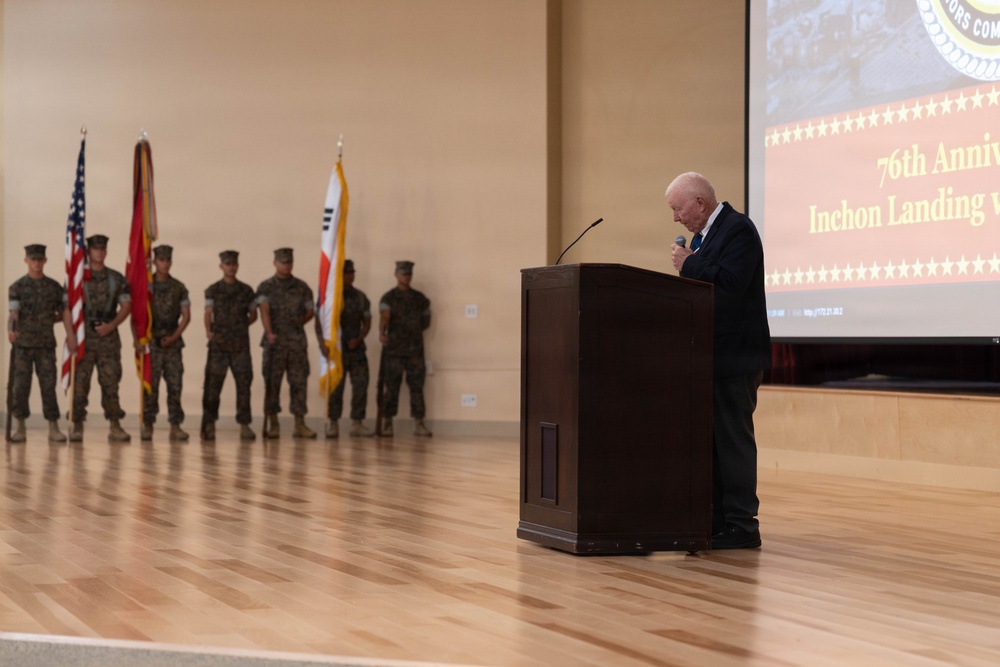A formal military ceremony commemorating the 76th Anniversary of the Inchon Landing is taking place in an indoor venue. An elderly civilian speaker addresses the audience from a wooden podium while a color guard of uniformed Marines stands at attention in the background. Flags including the United States flag, a Marine Corps flag, and what appears to be the South Korean flag are displayed.