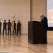 A formal military ceremony commemorating the 76th Anniversary of the Inchon Landing is taking place in an indoor venue. An elderly civilian speaker addresses the audience from a wooden podium while a color guard of uniformed Marines stands at attention in the background. Flags including the United States flag, a Marine Corps flag, and what appears to be the South Korean flag are displayed.