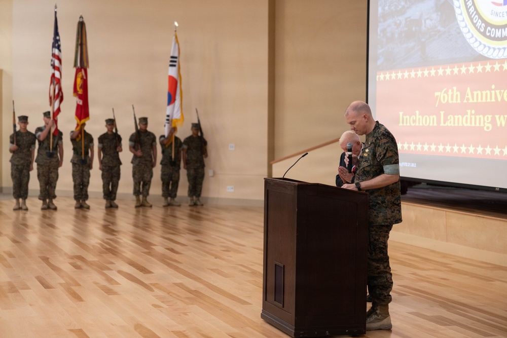 A military ceremony commemorating the 76th Anniversary of the Inchon Landing is taking place in a formal indoor venue. A Marine in camouflage uniform stands at a wooden podium embracing an elderly individual, likely a Korean War veteran. A color guard stands at attention in the background holding multiple flags including the U.S. flag, Marine Corps flag, and South Korean flag.