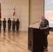 A military ceremony commemorating the 76th Anniversary of the Inchon Landing is taking place in a formal indoor venue. A Marine in camouflage uniform stands at a wooden podium embracing an elderly individual, likely a Korean War veteran. A color guard stands at attention in the background holding multiple flags including the U.S. flag, Marine Corps flag, and South Korean flag.