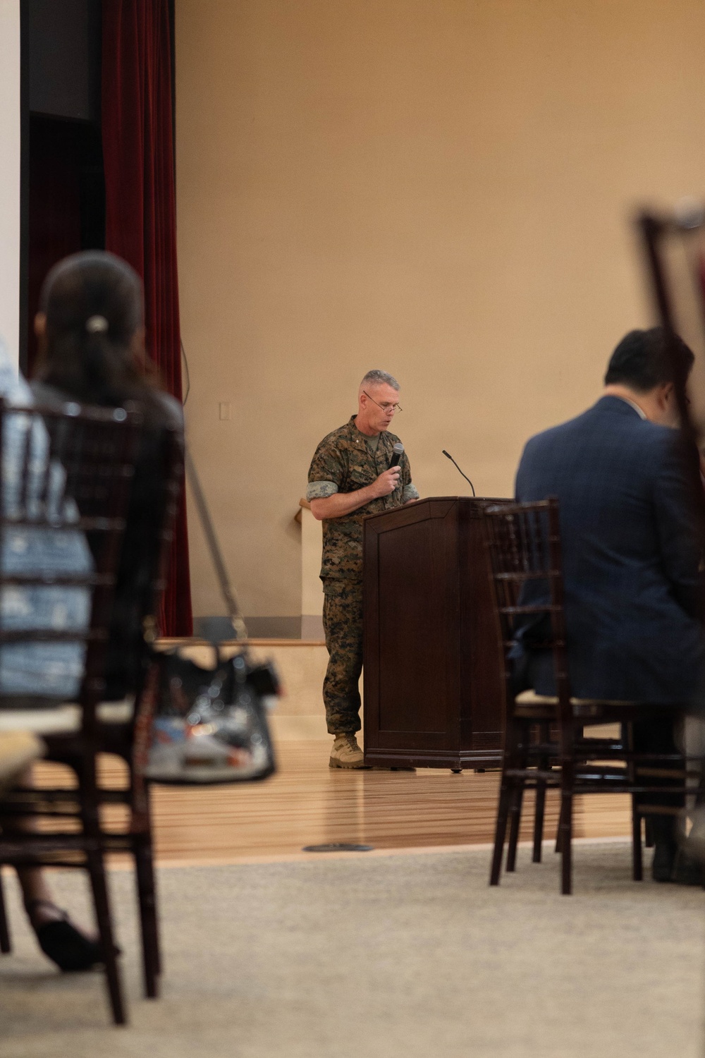 A military officer in USMC MARPAT camouflage uniform stands at a wooden podium, appearing to deliver a speech or briefing to an audience. The setting is an indoor hall or event space with beige walls and a raised stage area with red curtains. Audience members are visible in the foreground, seated in dark wooden chairs.