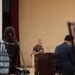 A military officer in USMC MARPAT camouflage uniform stands at a wooden podium, appearing to deliver a speech or briefing to an audience. The setting is an indoor hall or event space with beige walls and a raised stage area with red curtains. Audience members are visible in the foreground, seated in dark wooden chairs.