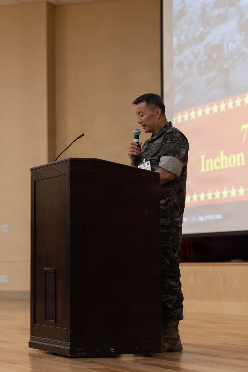 A military officer in camouflage uniform stands at a wooden podium holding a handheld microphone, delivering a speech or presentation. Behind him, a projection screen displays a slide referencing 'Inchon' with stars and what appears to be a historical military photograph, suggesting a commemorative or educational event. The setting appears to be a formal auditorium or conference hall.