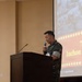 A military officer in camouflage uniform stands at a wooden podium holding a handheld microphone, delivering a speech or presentation. Behind him, a projection screen displays a slide referencing 'Inchon' with stars and what appears to be a historical military photograph, suggesting a commemorative or educational event. The setting appears to be a formal auditorium or conference hall.