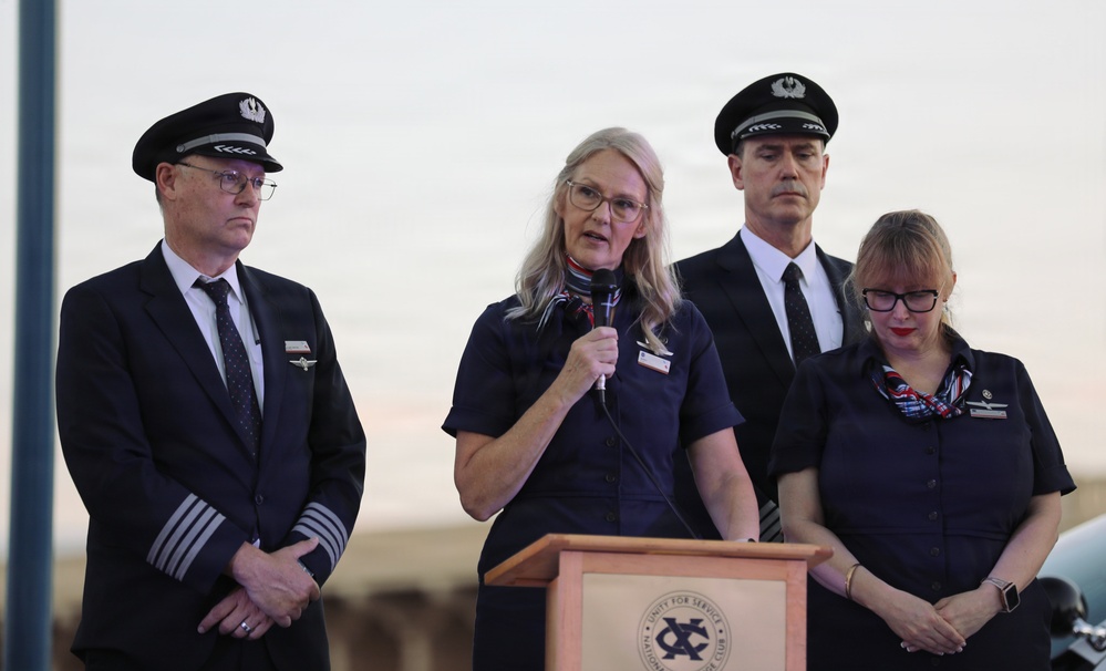 Four airline crew members are gathered at a podium during what appears to be a formal outdoor ceremony or press event. A woman in a navy flight attendant uniform is speaking into a microphone at the lectern, flanked by two pilots in uniform caps and another flight attendant. The group's solemn expressions suggest this may be a memorial or serious official address.