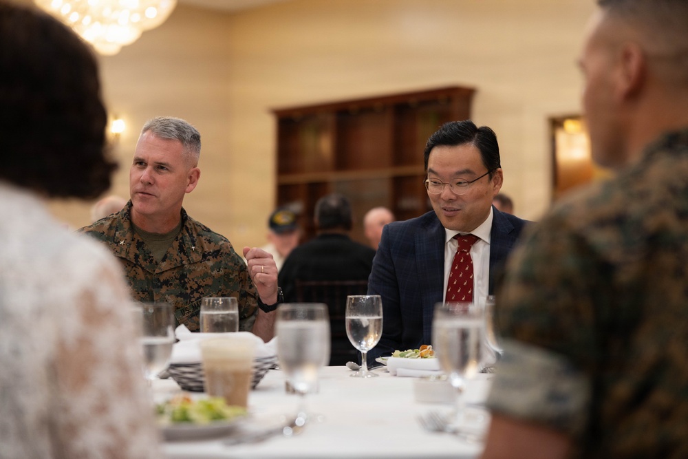 A formal dining event featuring military personnel in U.S. Marine Corps camouflage uniforms seated at a white-tablecloth table alongside civilians. The setting appears to be an official military dining facility or banquet hall, with multiple attendees engaged in conversation. The event has the appearance of a formal luncheon or dinner meeting between military and civilian officials.