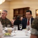 A formal dining event featuring military personnel in U.S. Marine Corps camouflage uniforms seated at a white-tablecloth table alongside civilians. The setting appears to be an official military dining facility or banquet hall, with multiple attendees engaged in conversation. The event has the appearance of a formal luncheon or dinner meeting between military and civilian officials.