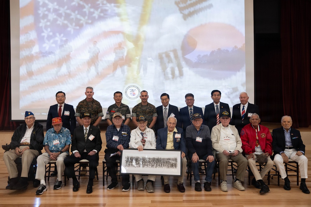 A formal group photograph of Korean War veterans seated in the front row, accompanied by military personnel and officials standing behind them. Two veterans in the center are holding a framed black-and-white panoramic photograph, likely a historical military unit photo. The event appears to be a commemorative ceremony, with a projected backdrop displaying the American and South Korean flags along with an organizational seal.