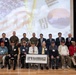 A formal group photograph of Korean War veterans seated in the front row, accompanied by military personnel and officials standing behind them. Two veterans in the center are holding a framed black-and-white panoramic photograph, likely a historical military unit photo. The event appears to be a commemorative ceremony, with a projected backdrop displaying the American and South Korean flags along with an organizational seal.