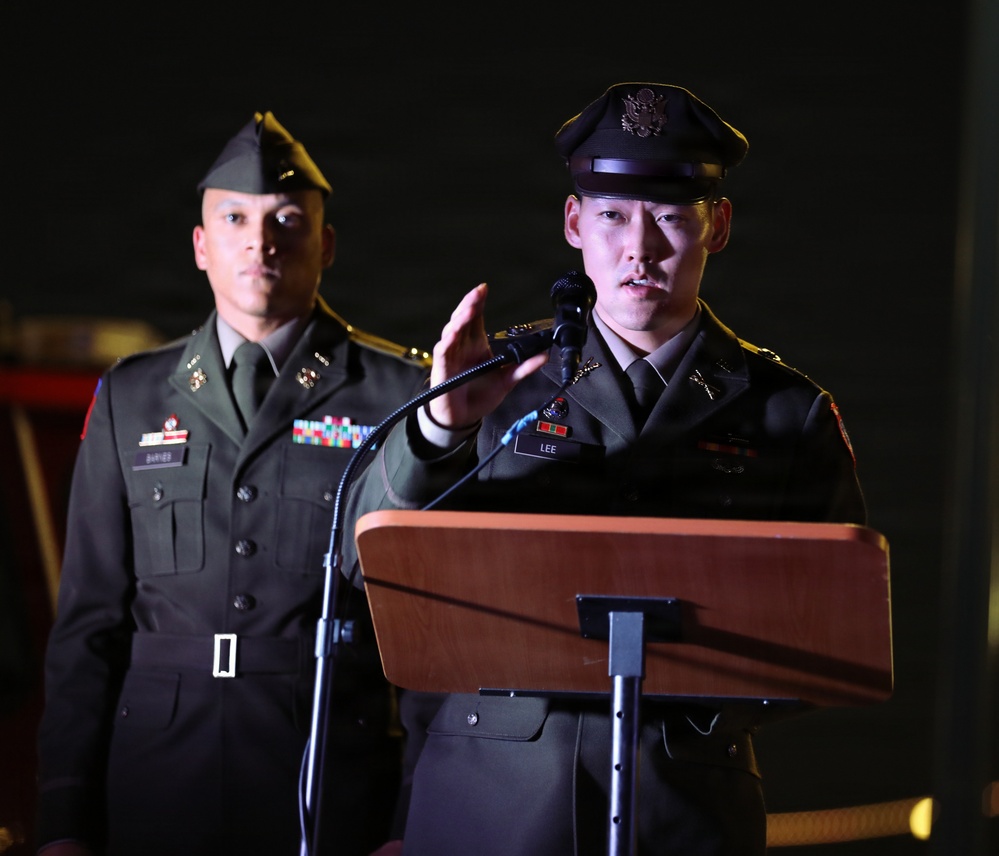 Two U.S. Army soldiers in dress uniforms are participating in a formal military ceremony or event. The soldier in the foreground, identified by a name tag reading 'LEE,' is speaking at a podium with a microphone. A second soldier stands at attention in the background.