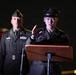 Two U.S. Army soldiers in dress uniforms are participating in a formal military ceremony or event. The soldier in the foreground, identified by a name tag reading 'LEE,' is speaking at a podium with a microphone. A second soldier stands at attention in the background.