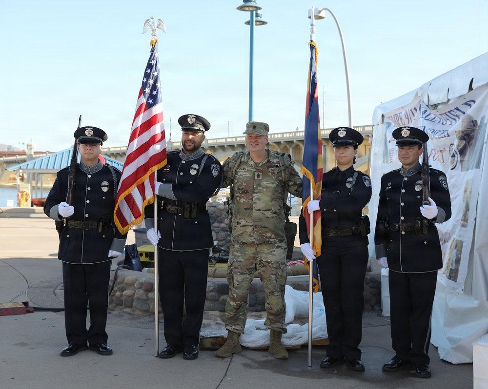 A color guard ceremony featuring four police officers in formal dark uniforms and one military personnel in camouflage (OCP/MultiCam) uniform posing together outdoors. The group is holding the United States flag and what appears to be the Arizona state flag, suggesting this is an official ceremonial event. The setting appears to be near a waterfront or marina area with a bridge visible in the background.