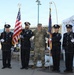 A color guard ceremony featuring four police officers in formal dark uniforms and one military personnel in camouflage (OCP/MultiCam) uniform posing together outdoors. The group is holding the United States flag and what appears to be the Arizona state flag, suggesting this is an official ceremonial event. The setting appears to be near a waterfront or marina area with a bridge visible in the background.