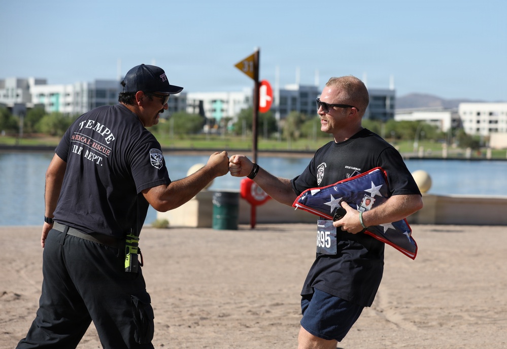 Two men wearing fire department shirts are fist-bumping near a waterfront area. The man on the right, wearing bib number 895 and identified as 'Kyle Carman' on his shirt, is carrying a folded American flag with what appears to be a badge or photo attached. This appears to be a memorial run or first responder event near Tempe, Arizona.