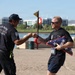 Two men wearing fire department shirts are fist-bumping near a waterfront area. The man on the right, wearing bib number 895 and identified as 'Kyle Carman' on his shirt, is carrying a folded American flag with what appears to be a badge or photo attached. This appears to be a memorial run or first responder event near Tempe, Arizona.