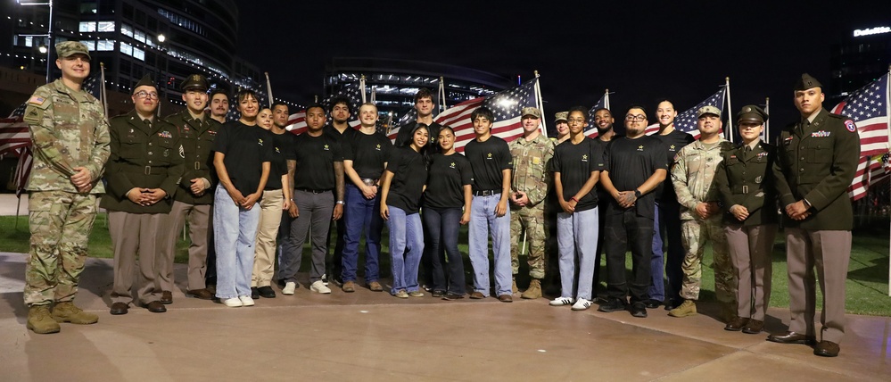 A group of approximately 25 individuals pose for a formal group photo at night in front of multiple American flags. The group includes both military personnel in uniform (Army combat uniforms and dress greens) and civilians wearing matching black t-shirts with what appears to be a U.S. Army recruiting logo. The setting appears to be an outdoor plaza or public space near a large stadium or arena.