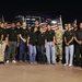 A group of approximately 25 individuals pose for a formal group photo at night in front of multiple American flags. The group includes both military personnel in uniform (Army combat uniforms and dress greens) and civilians wearing matching black t-shirts with what appears to be a U.S. Army recruiting logo. The setting appears to be an outdoor plaza or public space near a large stadium or arena.