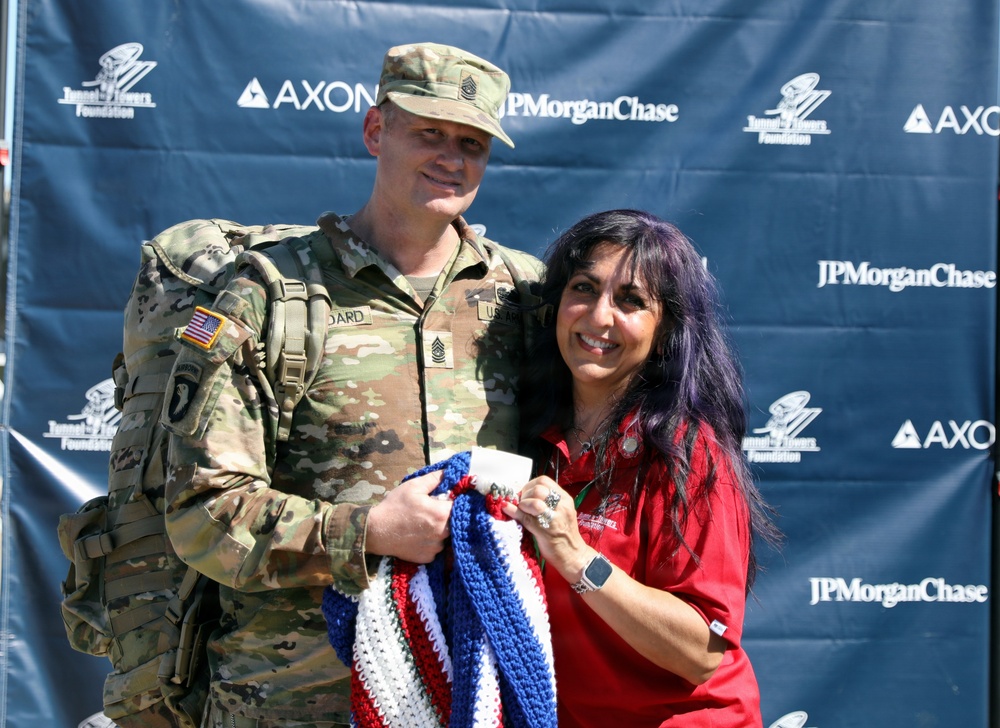 A U.S. Army soldier in full combat uniform and a woman in a red shirt pose together in front of a sponsor backdrop. They are jointly holding a red, white, and blue crocheted/knitted blanket, suggesting a ceremonial presentation or donation. The backdrop features logos from Tunnel to Towers Foundation, AXON, and JPMorgan Chase.