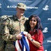 A U.S. Army soldier in full combat uniform and a woman in a red shirt pose together in front of a sponsor backdrop. They are jointly holding a red, white, and blue crocheted/knitted blanket, suggesting a ceremonial presentation or donation. The backdrop features logos from Tunnel to Towers Foundation, AXON, and JPMorgan Chase.