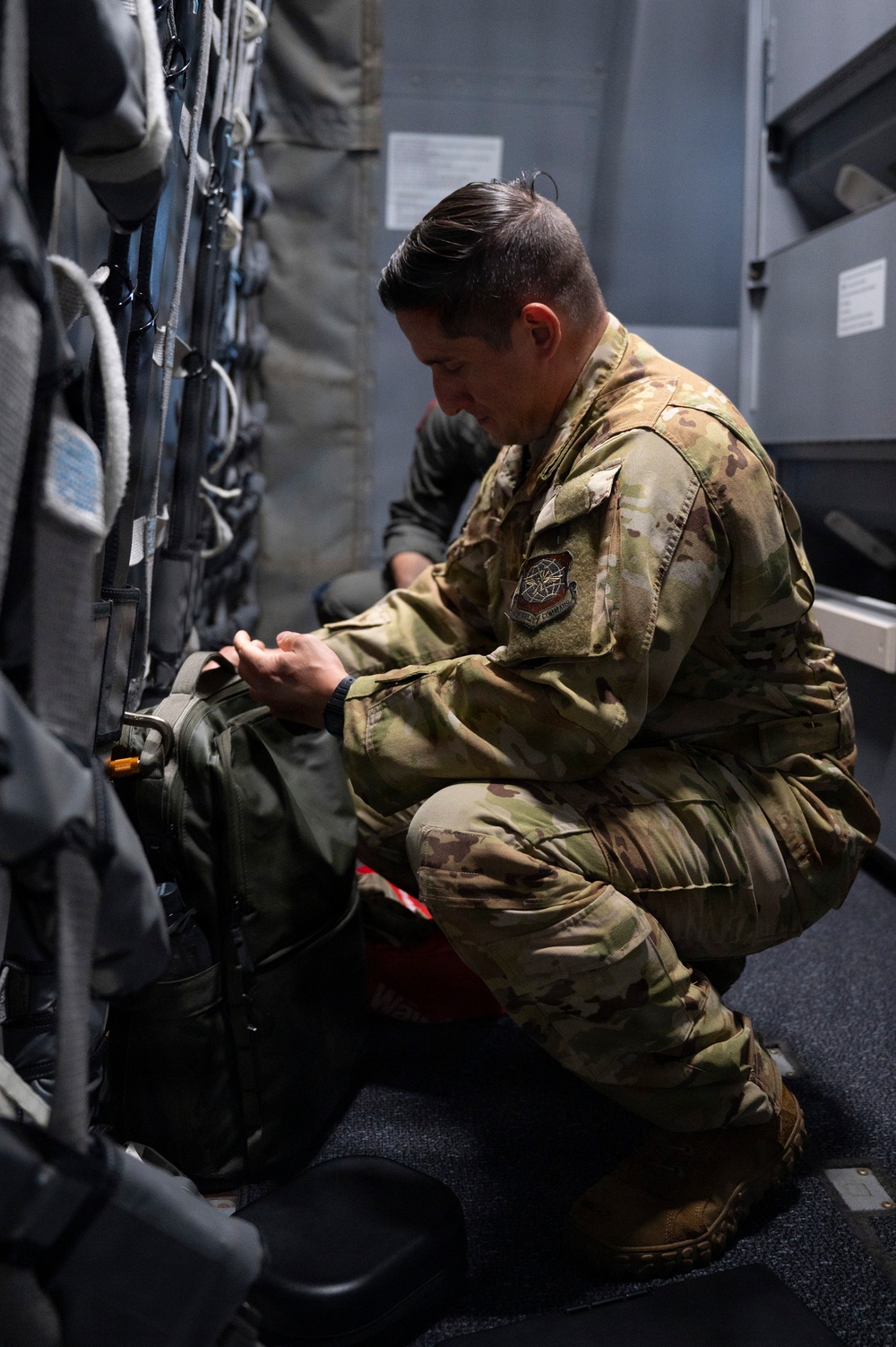 A military service member in OCP (Operational Camouflage Pattern) uniform is crouched down inspecting or adjusting equipment inside what appears to be a military aircraft. The individual is wearing a unit patch on their left shoulder and a watch, and is handling what appears to be a green military bag or pack secured to the aircraft wall. The interior setting shows straps, harnesses, and storage compartments typical of a military transport aircraft.
