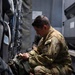 A military service member in OCP (Operational Camouflage Pattern) uniform is crouched down inspecting or adjusting equipment inside what appears to be a military aircraft. The individual is wearing a unit patch on their left shoulder and a watch, and is handling what appears to be a green military bag or pack secured to the aircraft wall. The interior setting shows straps, harnesses, and storage compartments typical of a military transport aircraft.