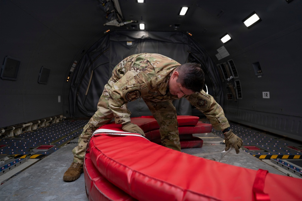 A military service member in OCP (Operational Camouflage Pattern) uniform is working inside the cargo hold of a large military transport aircraft, handling what appears to be red inflatable equipment or padding. The interior shows the characteristic ribbed walls, roller-equipped floor tracks, and overhead lighting of a wide-body military cargo aircraft, likely a C-17 or similar platform. The service member appears to be securing or arranging the red equipment on the aircraft floor.