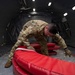 A military service member in OCP (Operational Camouflage Pattern) uniform is working inside the cargo hold of a large military transport aircraft, handling what appears to be red inflatable equipment or padding. The interior shows the characteristic ribbed walls, roller-equipped floor tracks, and overhead lighting of a wide-body military cargo aircraft, likely a C-17 or similar platform. The service member appears to be securing or arranging the red equipment on the aircraft floor.