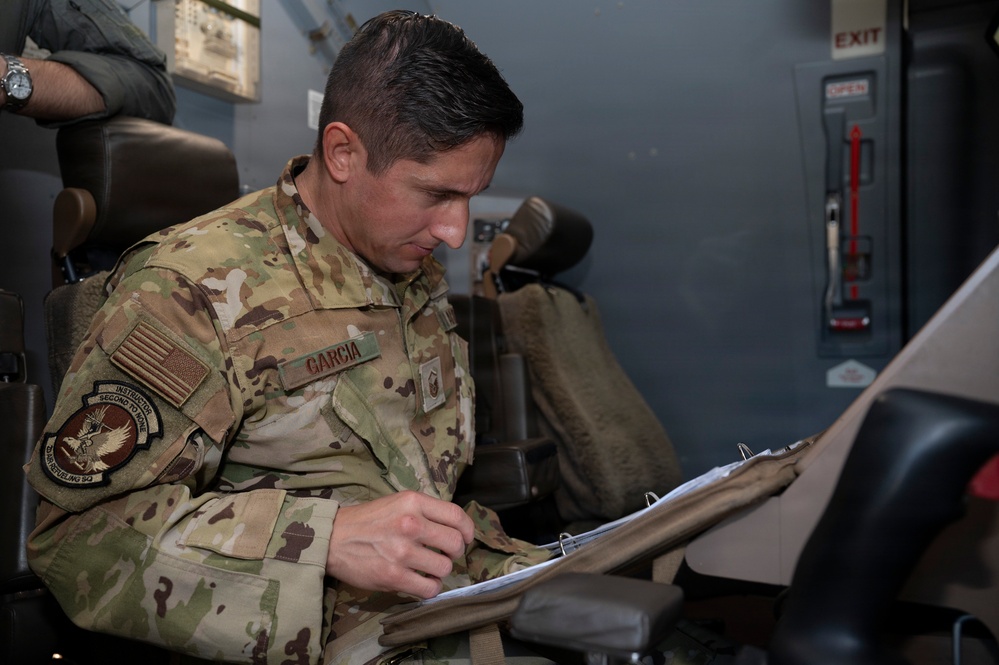 A U.S. Air Force service member named Garcia, wearing OCP (Operational Camouflage Pattern) uniform, is seated and reviewing documents in a binder. He has an instructor patch and a unit patch visible on his left sleeve, along with a U.S. flag patch. The setting appears to be inside a military aircraft or simulator environment.