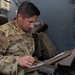 A U.S. Air Force service member named Garcia, wearing OCP (Operational Camouflage Pattern) uniform, is seated and reviewing documents in a binder. He has an instructor patch and a unit patch visible on his left sleeve, along with a U.S. flag patch. The setting appears to be inside a military aircraft or simulator environment.