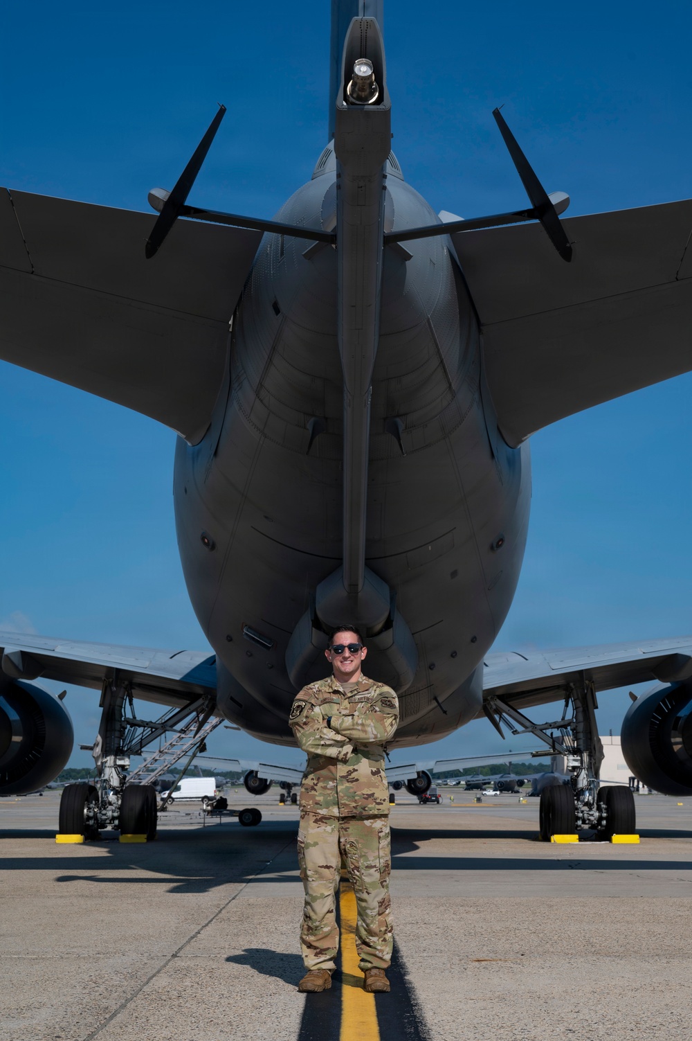 A military service member in OCP (Operational Camouflage Pattern) uniform stands with arms crossed on a flight line directly in front of a large military aircraft, likely a KC-46 Pegasus or similar tanker/transport aircraft. The photo is taken from a low angle looking up toward the nose of the aircraft, emphasizing its massive scale relative to the individual. The setting appears to be a military air base on a clear, sunny day.