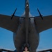 A military service member in OCP (Operational Camouflage Pattern) uniform stands with arms crossed on a flight line directly in front of a large military aircraft, likely a KC-46 Pegasus or similar tanker/transport aircraft. The photo is taken from a low angle looking up toward the nose of the aircraft, emphasizing its massive scale relative to the individual. The setting appears to be a military air base on a clear, sunny day.