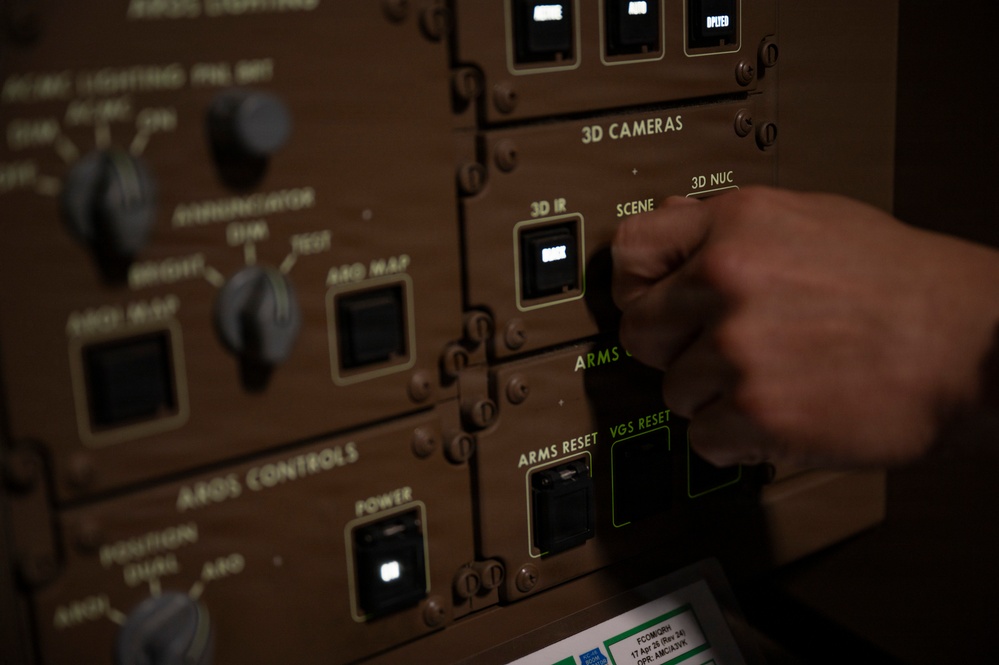 A hand is interacting with a military aircraft control panel, specifically in the area labeled '3D CAMERAS' with subsections marked '3D IR', 'SCENE', and '3D NUC'. The panel is olive/tan colored, consistent with military avionics equipment, and features illuminated switches and labeled controls. A document or placard is partially visible at the bottom of the frame, dated '17 Apr 24'.