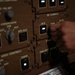 A hand is interacting with a military aircraft control panel, specifically in the area labeled '3D CAMERAS' with subsections marked '3D IR', 'SCENE', and '3D NUC'. The panel is olive/tan colored, consistent with military avionics equipment, and features illuminated switches and labeled controls. A document or placard is partially visible at the bottom of the frame, dated '17 Apr 24'.