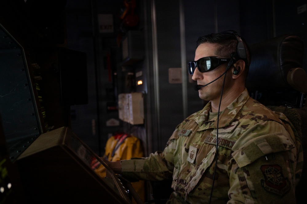 A U.S. Air Force service member is seated at a control station inside what appears to be a military aircraft. The individual is wearing OCP (Operational Camouflage Pattern) uniform, tactical sunglasses, and a communications headset with a boom microphone. The setting suggests an active operational or in-flight environment.