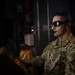 A U.S. Air Force service member is seated at a control station inside what appears to be a military aircraft. The individual is wearing OCP (Operational Camouflage Pattern) uniform, tactical sunglasses, and a communications headset with a boom microphone. The setting suggests an active operational or in-flight environment.
