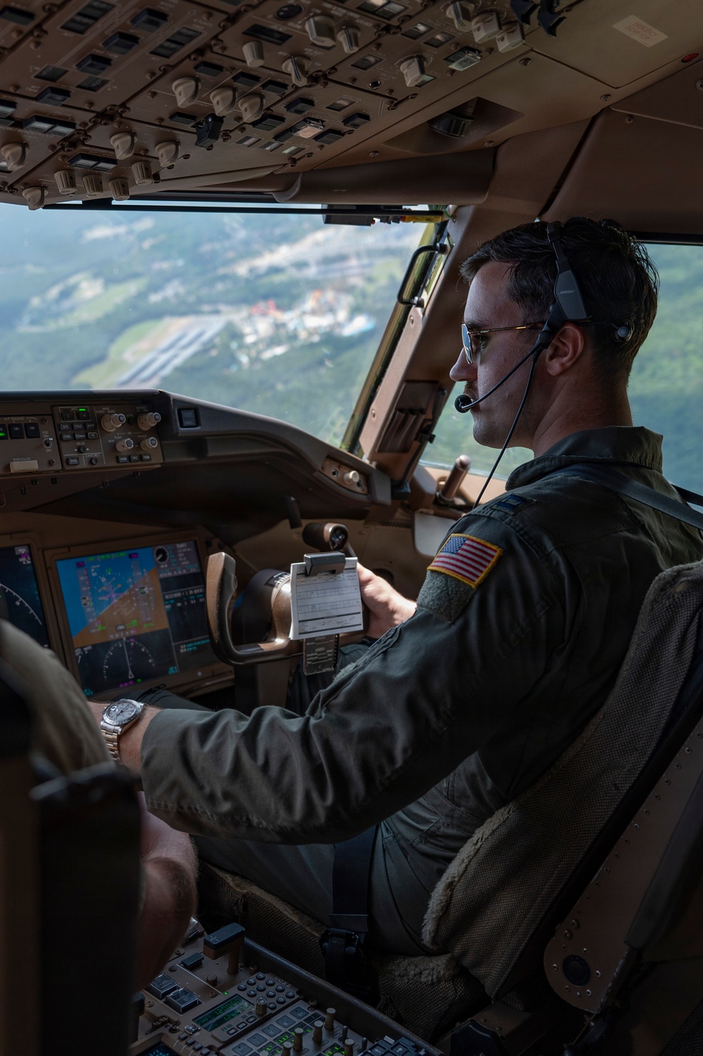 A U.S. military pilot is seated in the cockpit of a large military transport aircraft, actively flying at low-to-medium altitude over a green, hilly landscape with visible infrastructure below. He is wearing an olive drab flight suit with a U.S. flag patch on the right shoulder, aviator sunglasses, and a Bose aviation headset with boom microphone. The cockpit features advanced glass avionics displays and a complex overhead panel consistent with a Boeing C-17 Globemaster III.