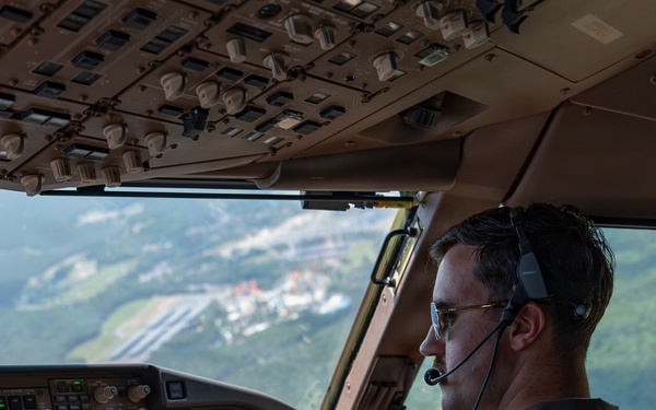 A U.S. military pilot is seated in the cockpit of a large military transport aircraft, actively flying at low-to-medium altitude over a green, hilly landscape with visible infrastructure below. He is wearing an olive drab flight suit with a U.S. flag patch on the right shoulder, aviator sunglasses, and a Bose aviation headset with boom microphone. The cockpit features advanced glass avionics displays and a complex overhead panel consistent with a Boeing C-17 Globemaster III.