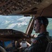A U.S. military pilot is seated in the cockpit of a large military transport aircraft, actively flying at low-to-medium altitude over a green, hilly landscape with visible infrastructure below. He is wearing an olive drab flight suit with a U.S. flag patch on the right shoulder, aviator sunglasses, and a Bose aviation headset with boom microphone. The cockpit features advanced glass avionics displays and a complex overhead panel consistent with a Boeing C-17 Globemaster III.