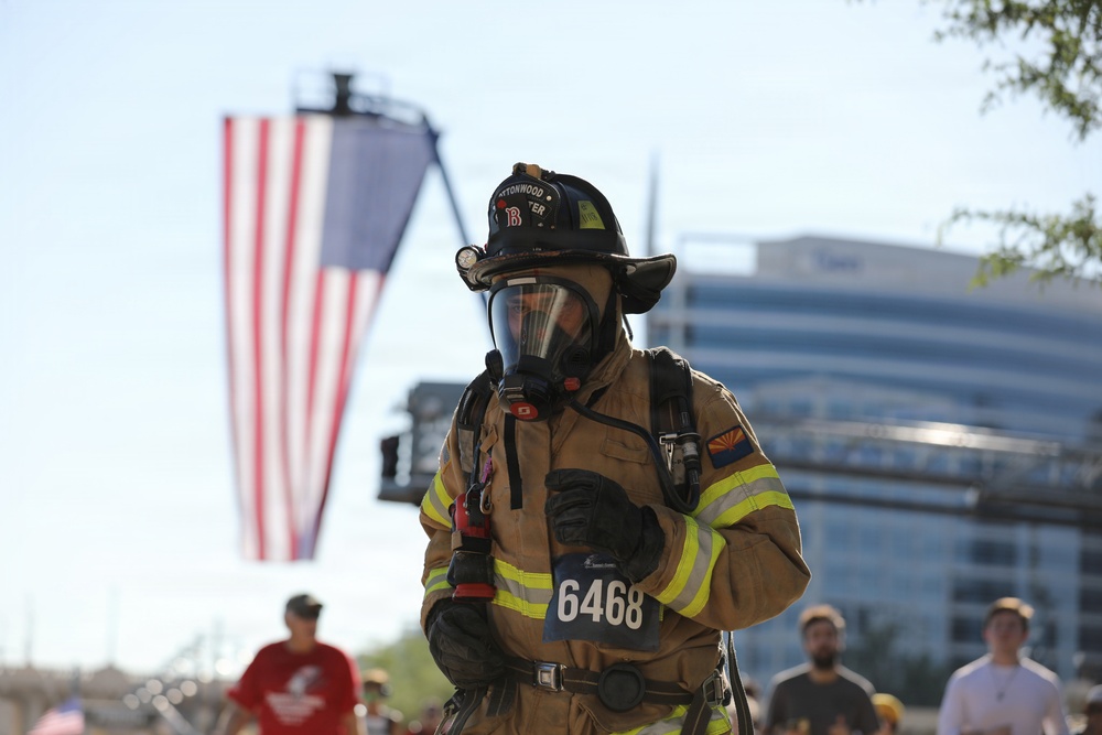 A firefighter in full turnout gear, including SCBA mask and helmet, participates in what appears to be a memorial run or charity race, indicated by the race bib number 6468. A large American flag is displayed in the background, suggesting a patriotic or commemorative event. The setting appears to be an urban outdoor location with other participants and a modern building visible in the background.