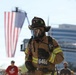 A firefighter in full turnout gear, including SCBA mask and helmet, participates in what appears to be a memorial run or charity race, indicated by the race bib number 6468. A large American flag is displayed in the background, suggesting a patriotic or commemorative event. The setting appears to be an urban outdoor location with other participants and a modern building visible in the background.